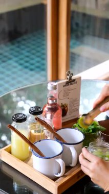 Bakire mojito hazırlarken çekilmiş dikey bir video. Kadın elleri, tarçın çubuğuyla limon dilimlerini bardağa koyar. Arka planda havuz manzaralı tropikal bir villada içecek hazırlıyorum.