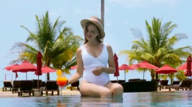 Young woman in white swimsuit drinks red orange non-alcoholic cocktail, relax sitting in swimming pool water. Smiling female tourist in hat enjoying vacation at luxury hotel or resort in summer heat.