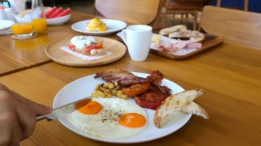 Masa bıçağıyla erkek eli, yumurtanın güneşli tarafını kesmiş, yumurtanın içi sıvı sarısı ile dolu, restoranda beyaz tabakta. Kahvaltı, öğle yemeği ya da brunch yemeği. Adam kızarmış ekmek, domuz pastırması ve domatesle kafede kızarmış yumurta yiyor.