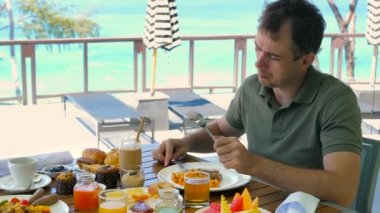 Young man have breakfast or brunch at luxury restaurant in hotel with turquoise sea background. Male tourist drinking coffee at morning in resort cafe. Wanderlust, travel, holiday, vacation concept.