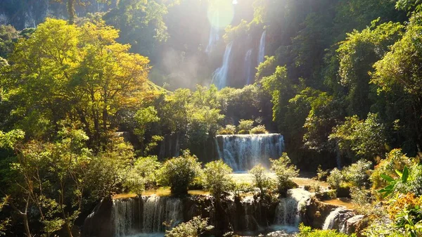 Beautiful waterfall in green tropical forest. Mountain jungle with limestone waterfalls cascades. Powerful raging whitewater falling stream. Slow motion. Lens flare, shining sun, hot summer day.