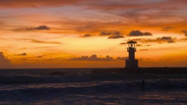 Scenic evening tropical seascape during sunset time, silhouettes of surfers on surfboards at sea waves, orange sky, flying birds, lighthouse. Wanderlust, summer travel, holidays, vacations concept.