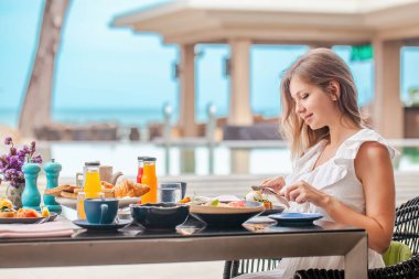 Lüks tropikal tatil köyünde, havuz kenarında kahvaltı yapan bir kadın. Çeşitli yiyecek ve içeceklerle dolu bir masa. Sağlıklı kahvaltı ve meyve tabakları. Plaj lüks yaşam tarzı, yaz tatili.