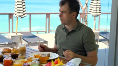 Young man eating brunch at restaurant in tropical hotel with turquoise sea background. Serious male tourist have breakfast at morning in resort cafe. Wanderlust, travel, holiday, vacation concept.