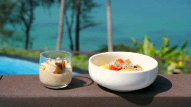 Healthy breakfast food on ocean beach background. Oatmeal in jars with almond petals and bowl with fresh tropical fruits. Breakfast in a beautiful summer location with sea views. Vegan eating.