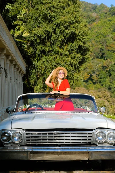 Young woman enjoying road trip on the ancient columns and palm trees on background, standing in convertible car. Stylish traveller girl in red dress and hat on vacation. Vertical video