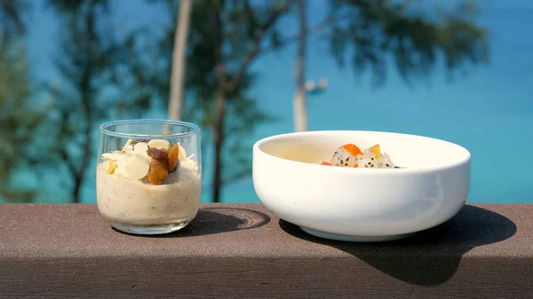Healthy breakfast food on ocean beach background. Oatmeal in jars with almond petals and bowl with fresh tropical fruits. Breakfast in a beautiful summer location with sea views. Vegan eating.