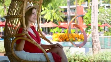 Romantic travel girl in straw hat and red dress rest at poolside in tropical resort on holidays. Attractive female tourist relaxing in rattan cocoon swing chair at luxury hotel in summer vacations.