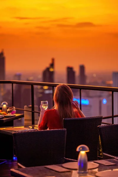 Young woman with glass of wine rest at luxury rooftop restaurant watching orange sky sunset. Female with cocktail drink at sky bar terrace looking at modern city skyline. Skyscrapers on background.