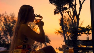 Young lady drinks cocktail sits in luxury outdoors bar or restaurant at hotel at evening sunset with sea and beautiful orange sky background. Cute woman in evening dress holds mocktail. Summer travel.