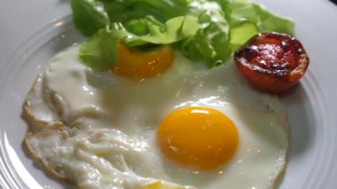 Concept of simple fresh healthy food on breakfast at home, fried eggs with tomato and salad leaf. Human hands with fork and knife cut sunny side up egg on white plate. Liquid egg yolk close-up.