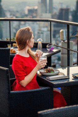 Kokteyl içen genç ve güzel bir kadın gün batımında lüks bir çatı restoranında dinleniyor. Gökyüzü bar terasında içkili kırmızı elbiseli zarif bir kadın modern şehir siluetine bakıyor. Arkaplanda gökdelenler.