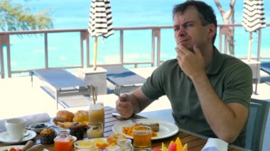 Sad thoughtful man touches face, touch chin by hand, have breakfast at restaurant in hotel, turquoise sea background. Unhappy depressed male tourist sighs. Bad, negative holiday, day off concept.