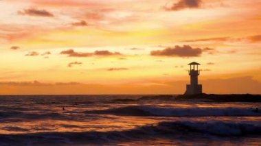 Scenic view of surf people sitting with surfboards, waiting ocean waves on sunset. Tropical beach with lighthouse on background. Concept of summer travel, adventure and extreme