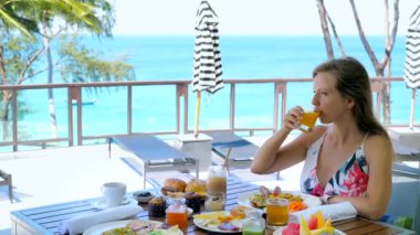 Happy smiling girl drinking orange juice on holiday brunch sitting by table full of food and drinks, pastry, coffee in sunny summer day, beautiful blue sea background. Travel, vacation concept.