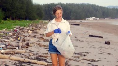 Kameraya bakan ve çöp torbasında plaj çöpü toplayan gönüllü bir kadın. Okyanus ve gezegen kirliliği kavramı. Çöp torbası olan kadın plajı plastik şişeden temizliyor.