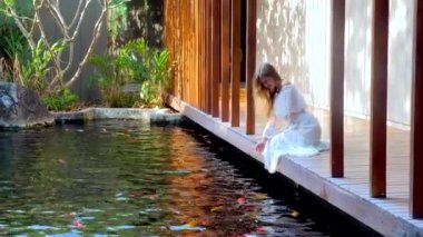 Japon bahçesinde balık besleyen bir kadın. Beyaz elbiseli kız tedavi sırasında tahta köprüde oturuyor, göl kenarında sazan balığıyla rahatlıyor. Spa, rahatlama ve rahatlama kavramı 