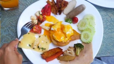 İnsan eli çatalla otel veya tatil köyündeki restoranda brunch yer. Dilimlenmiş Benedict yumurta sarısı, füme somon dilimi, hollandaise sosu, pastırma, sosis, peynir ve ızgara sebze ile kahvaltı tabağı..