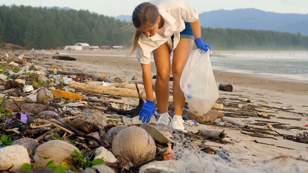 Plastic ocean pollution. Woman in gloves walking on the sea beach, picks up empty bottles trash and rubbish from the sand and collect in the trash bag. Volunteer girl help cleaning ocean coastline