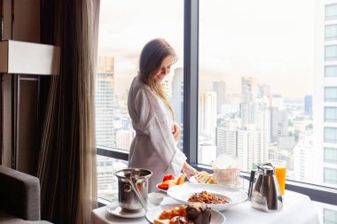 Room service in luxury hotel. Woman in bathrobe eating breakfast near window with city megapolis view. Served food on table in modern cozy room in hotel. Vacation or holidays concept