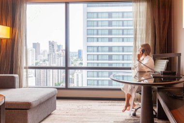 Bornozlu bir kadın modern bir otel ya da dairede oturuyor ve şehir gökdeleniyle telefonda konuşuyor. Lüks otellerde tatil. Otel odasında tatilde olan iş kadını.