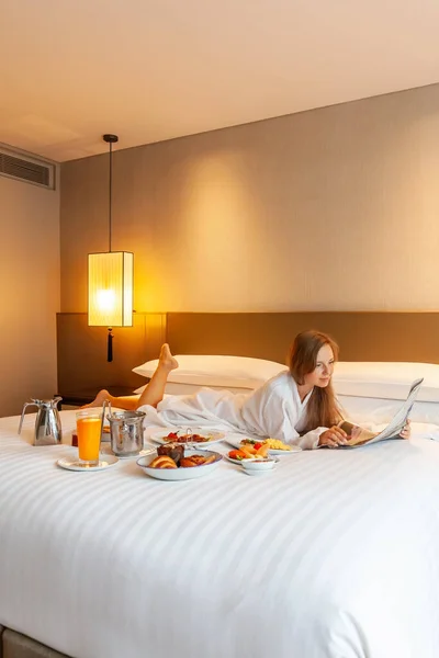 Food in bed serve in luxury hotel. Woman on vacation enjoy breakfast in bed in hotel room, reading newspaper, drinking orange juice, eating food and relaxing on holidays. Room Service