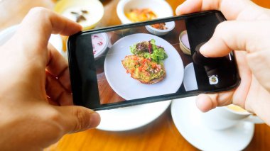 Erkek insan cep telefonuyla yemek fotoğrafı çeker. Adam elleri kafedeki ahşap masada sağlıklı kahvaltı vuruyor. Blogcu öğle yemeğini akıllı telefondan çekiyor. 