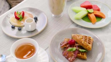 Belçika waffle 'ı, taze böğürtlenli krep, tropikal meyve, bir fincan sıcak çay ve buzlu kahve bardağı ile kafede tatlı kahvaltı. Lezzetli yemek, lezzetli tatlı. 