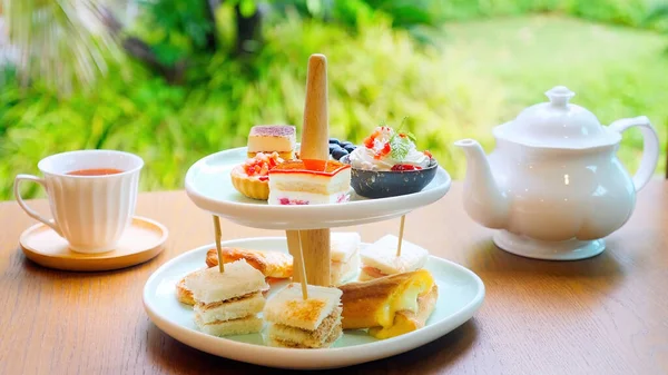 Tea time, traditional English tea ceremony with Afternoon tea cakes, sandwiches and teapot with hot tea. Sweet tasty cakes and desserts with greenery on background.