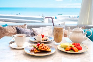 Belçika waffle 'ı, taze böğürtlenli krep, tropikal meyve, bir fincan sıcak çay ve buzlu kahve bardağı ile kafede tatlı kahvaltı. Lezzetli yemek, lezzetli tatlı. 