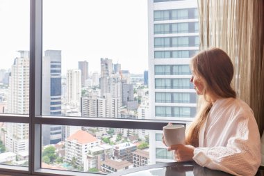 Modern bir otelde ya da gökdelen manzaralı bir dairede oturan bornozlu bir kadın. Lüks otellerde tatil. Tatilde otel odasında tanınmayan biri var.