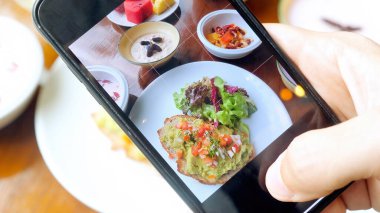 Erkek eller sağlıklı vejetaryen kahvaltısı ya da akşam yemeğini akıllı telefondan çekiyor. Erkek kişi cep telefonu tutuyor ve sosyal ağ için yemeğini çekiyor. Lokantadaki blog yazarı içerik oluşturur