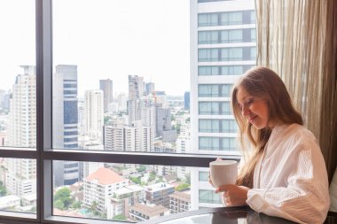 Modern bir otelde ya da gökdelen manzaralı bir dairede oturan bornozlu bir kadın. Lüks otellerde tatil. Otel odasında tatilde olan bir kadın.