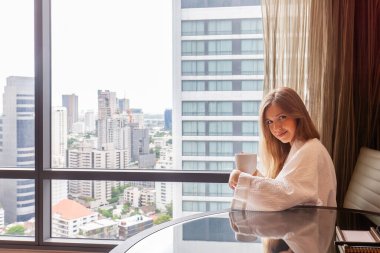 Modern bir otelde ya da gökdelen manzaralı bir dairede oturan bornozlu bir kadın. Lüks otellerde tatil. Otel odasında tatilde olan bir kadın.