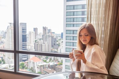 Modern bir otelde ya da gökdelen manzaralı bir dairede oturan bornozlu bir kadın. Lüks otellerde tatil. Otel odasında tatilde olan bir kadın.