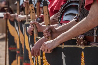 Romalı lejyonerler düzende. Pila ya da cirit tutan eller. Scuta üzerinde soluk kartal kanadı ve yıldırım motifleri