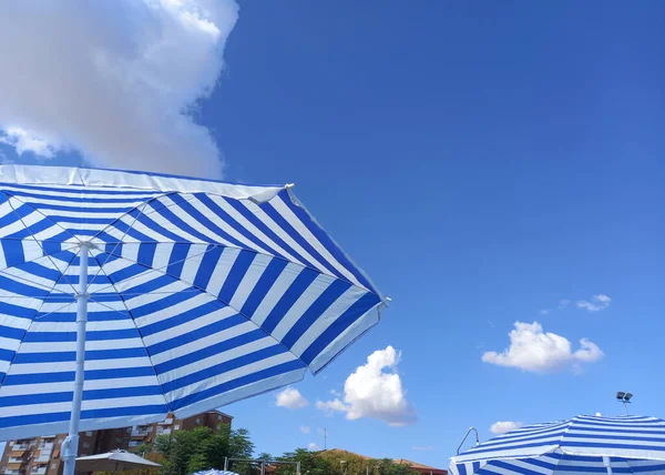 Blue striped umbrellas over blue sky with apartment building. Urban swimning pool background