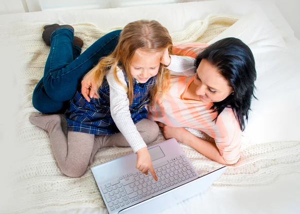 Happy mother and daughter working on computer sitting on bed together — Stock Photo, Image