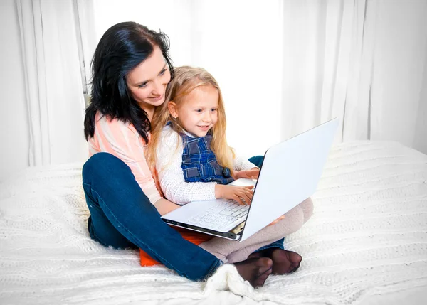 Happy mother and daughter working on computer sitting on bed together