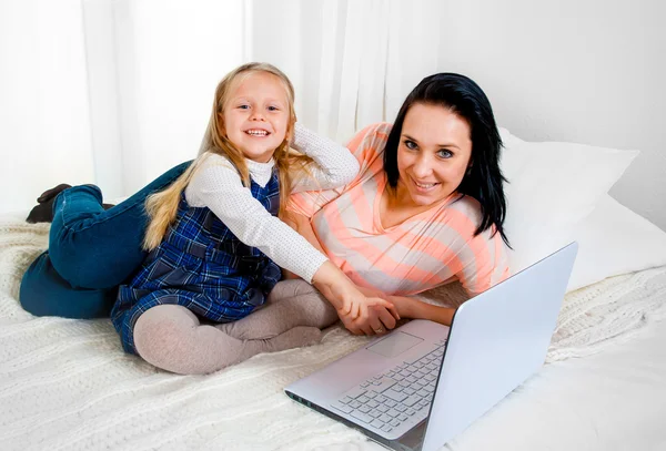 Happy mother and daughter working on computer sitting on bed together — Stock Photo, Image