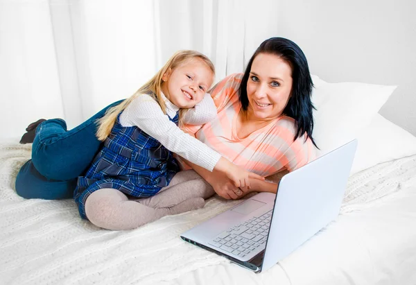 Happy mother and daughter working on computer sitting on bed together — Stock Photo, Image