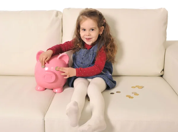 Милая маленькая девочка играет с монетами и огромной копилкой на диване — стоковое фото