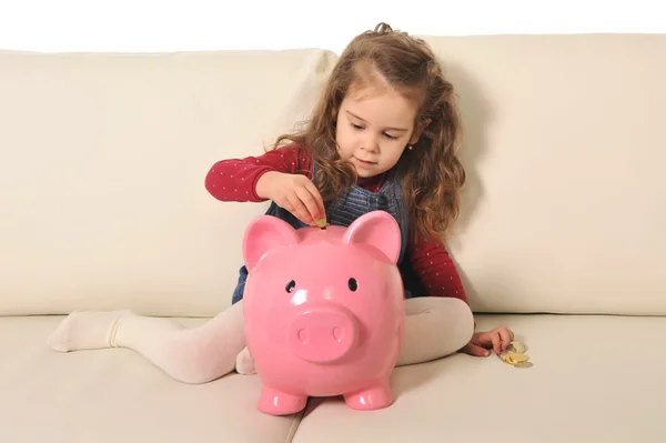 Nettes kleines Mädchen spielt legt Münze in riesiges Sparschwein auf Sofa — Stockfoto