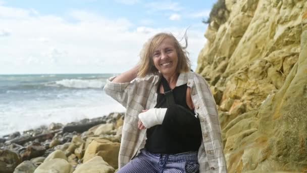 Senior anciana relajante sentada en la costa del océano o el mar ventoso día nublado que sufre de dolor. Lesión en brazo roto. Muñeca de señora mayor en yeso vendaje de honda sensación de dolor en el exterior en verano — Vídeos de Stock