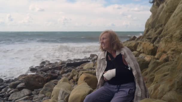 Mulher idosa sênior relaxante sentado na costa do oceano ou mar dia nublado ventoso sofrendo de dor. Lesão no braço partido. Pulso de senhora velha em gesso sling bandagem sentindo-se ferido fora no verão — Vídeo de Stock