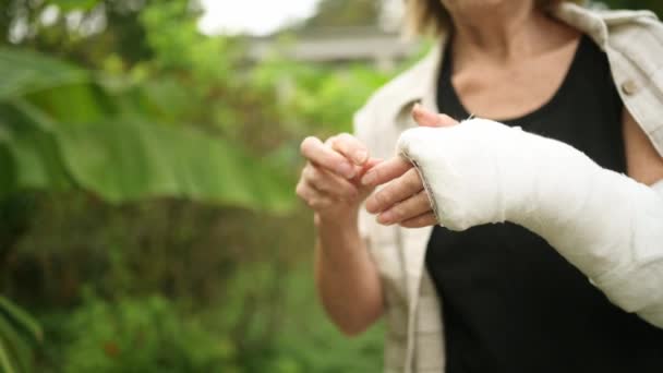 Idosa sênior massageando a mão sofrendo de dor nas articulações. Mulher mais velha com lesão braço quebrado no elenco fora no verão. Senhora mais velha tocando no pulso em gesso sling bandagem sentindo dor — Vídeo de Stock