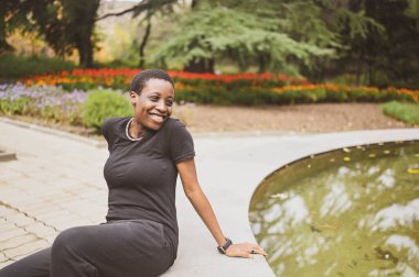 Çekici, mutlu, genç, doğal, güzel, kısa saçlı, siyah giyinmiş, yaz parkında bir çeşmenin kenarında oturan Afrikalı kadın..