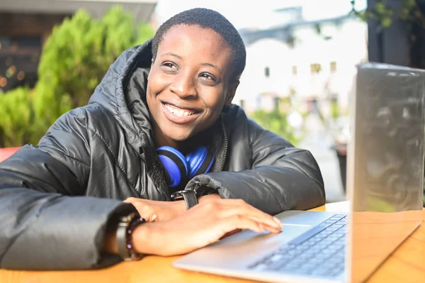Lachend vrolijke Afrikaanse zwarte kortharige studente in zwart donsjas met blauwe hoofdtelefoon die online werkt op een laptop computer in een café. Verscheidenheid. Afstandswerk, afstandsonderwijs. — Stockfoto