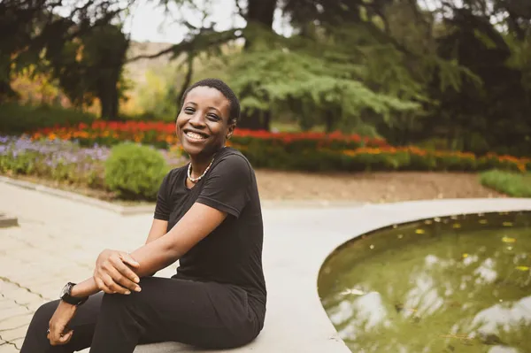 Menarik tersenyum muda kecantikan alam muda muda berambut pendek wanita Afrika mengenakan total hitam tertawa duduk di tepi air mancur di taman musim panas. — Stok Foto