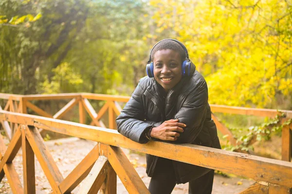 Attractive happy smile young natural beauty short haired African Black woman with blue headphone in black down jacket bersandar pada kayu railing listening music in nature summer park. — Stok Foto